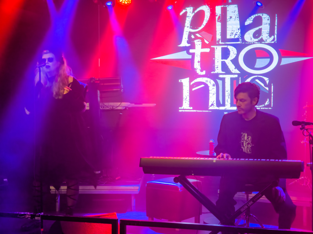 Platronic members on stage with blue and red stage light beams glowing through the haze. Vocalist Kay standing on the far left, producer Sami behind his keyboard on the right. Platronic logo projected on the wall behind.